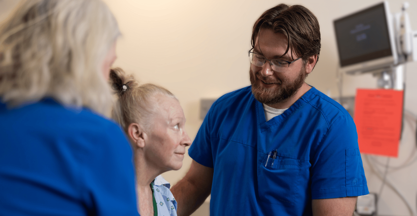 A healthcare professional in blue scrubs speaks with an older patient seated on an exam table in a clinical room.