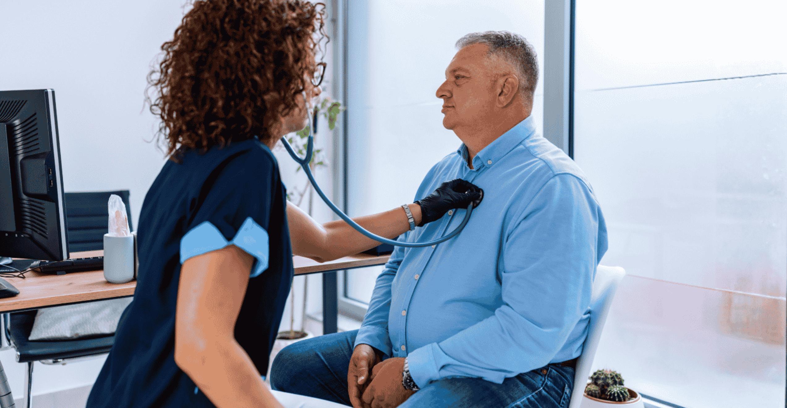 A healthcare professional wearing dark scrubs uses a stethoscope to listen to the chest of a seated adult patient in a light blue button-up shirt during a medical exam in a bright clinic room. A desk with a computer monitor and a tissue box is visible in the background, along with large windows letting in natural light.
