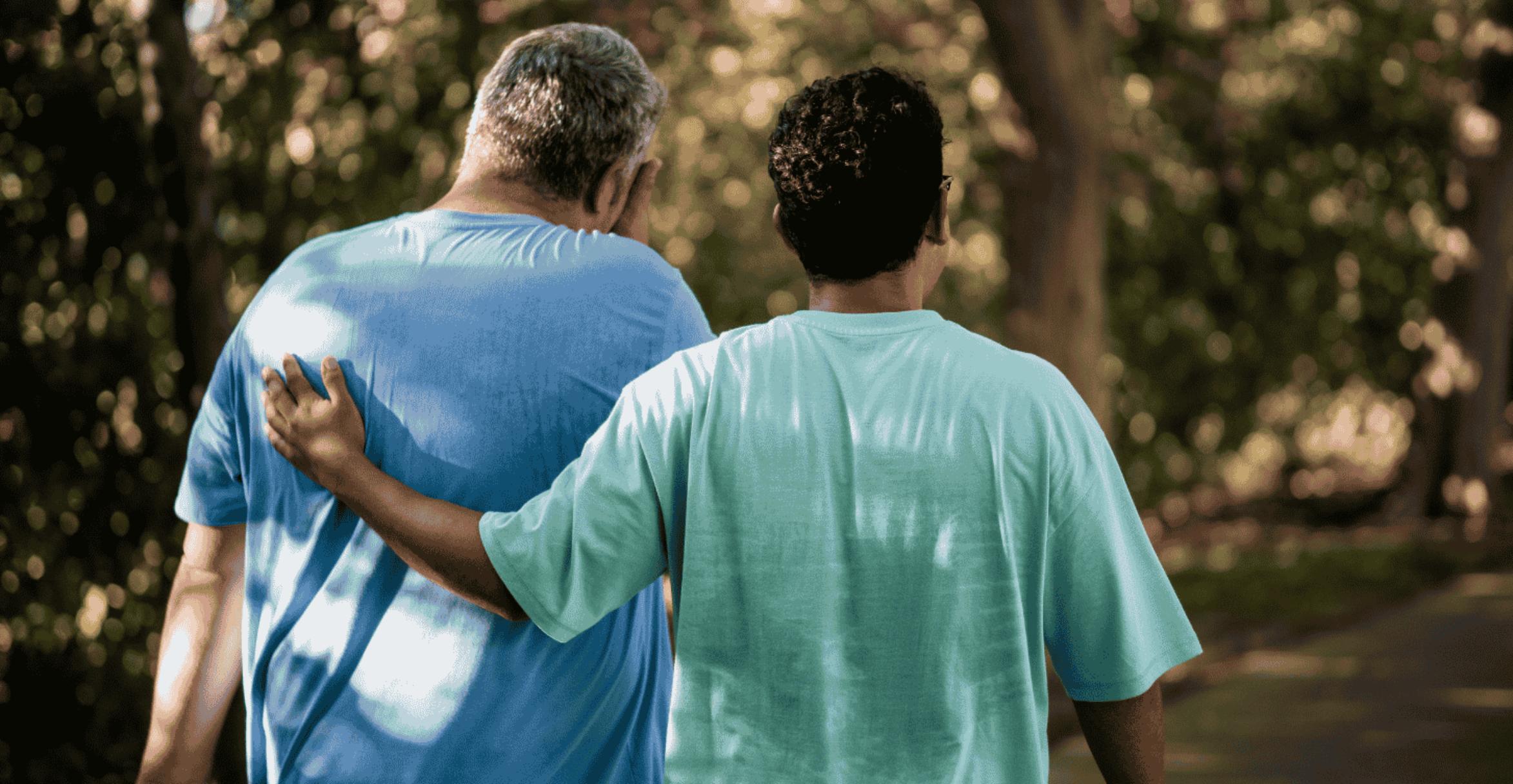 Two adults walk side by side along a tree-lined path, viewed from behind. One person wearing a light blue shirt places an arm around the shoulders of the other person wearing a light green shirt in a supportive gesture. Sunlight filters through the trees, casting soft, dappled light across the path and surrounding greenery.